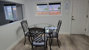 Un comedor con una mesa redonda de cristal y cuatro sillas, dos ventanas con vistas a edificios y montañas, y una puerta con un letrero.