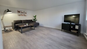 Una sala de estar moderna con un sofá gris, una mesa de centro, una lámpara de pie, un televisor sobre un soporte negro y una planta pequeña.