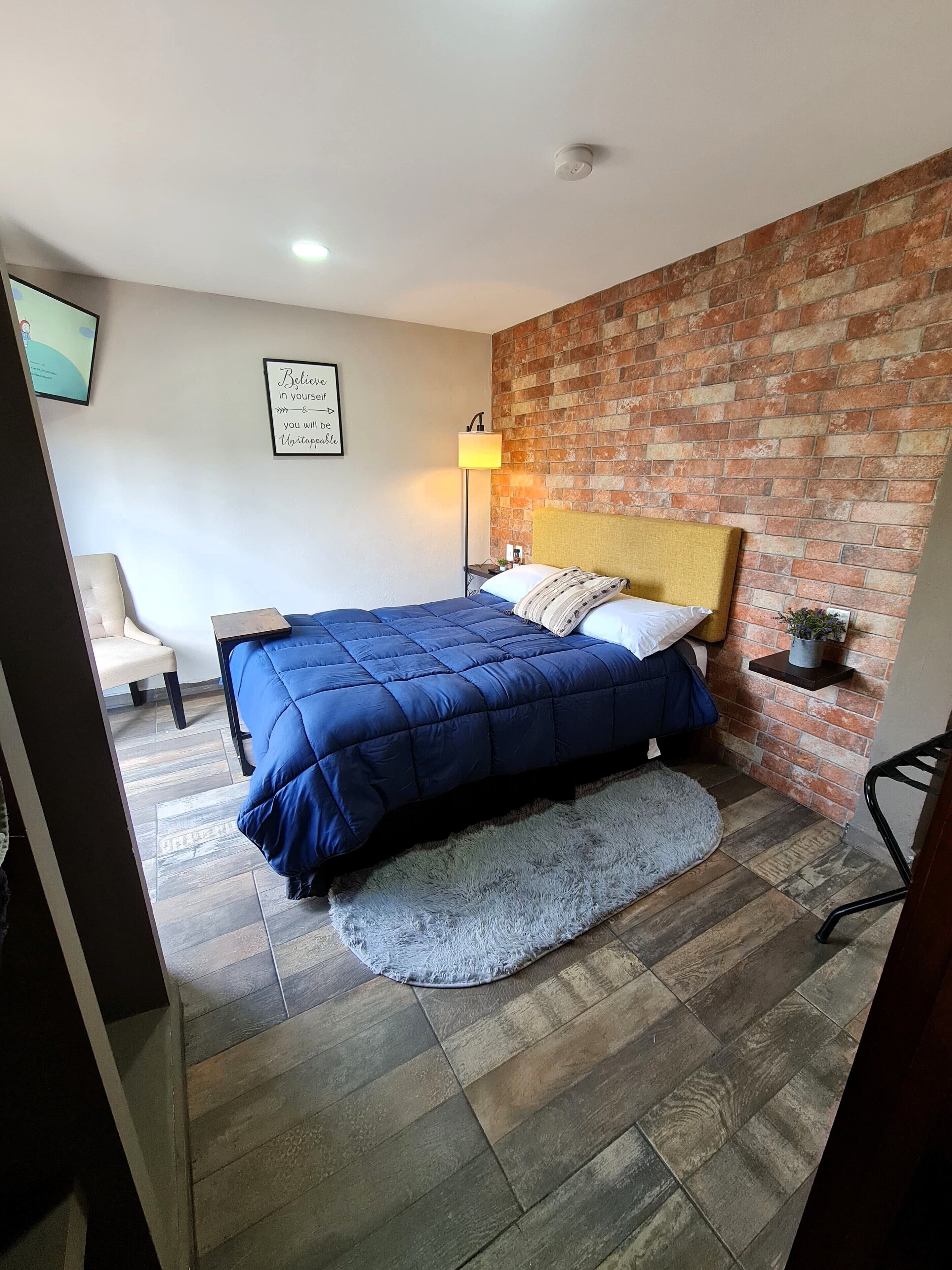 Un dormitorio con cama, mesita de noche, silla, estante en la pared y pared de ladrillo.