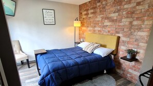 Un dormitorio con cama, mesita de noche, silla, estante en la pared y pared de ladrillo.