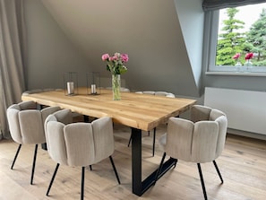 A dining area with a wooden table, six upholstered chairs, a vase with flowers, and a window with a view of trees.