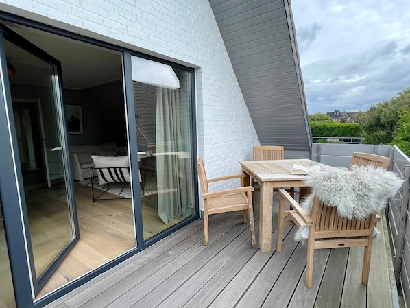 A modern outdoor patio with a wooden table and chairs, a glass door leading to an interior space, and a view of greenery in the background.