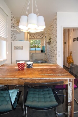 A dining area with a wooden table, two chairs with cushioned seats, a hanging light fixture, and a kitchen in the background. - Ferienhaus Westhüs - Modernes, Renoviertes Reihenendhaus in Tinnum mit 95qm für bis zu 6 Personen! (Sylt)