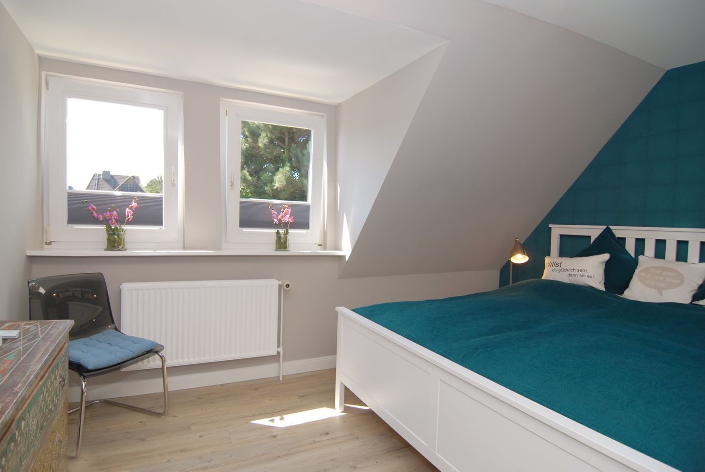 Une chambre à coucher avec un lit, une chaise, un bureau et deux fenêtres ornées de fleurs.