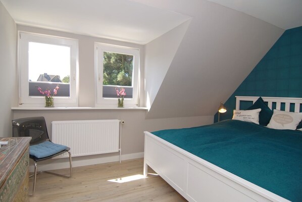 A bedroom with a bed, a chair, a desk, and two windows with flowers.