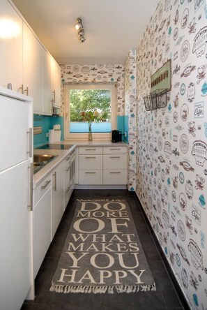 A narrow kitchen with white cabinets, a window with a view of greenery, and a wall with coffee-themed wallpaper. - Ferienhaus Westhüs - Modernes, Renoviertes Reihenendhaus in Tinnum mit 95qm für bis zu 6 Personen! (Sylt)