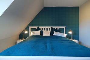 A bed with a white headboard, multiple pillows, and two bedside lamps in a room with a sloped ceiling. - Ferienhaus Westhüs - Modernes, Renoviertes Reihenendhaus in Tinnum mit 95qm für bis zu 6 Personen! (Sylt)