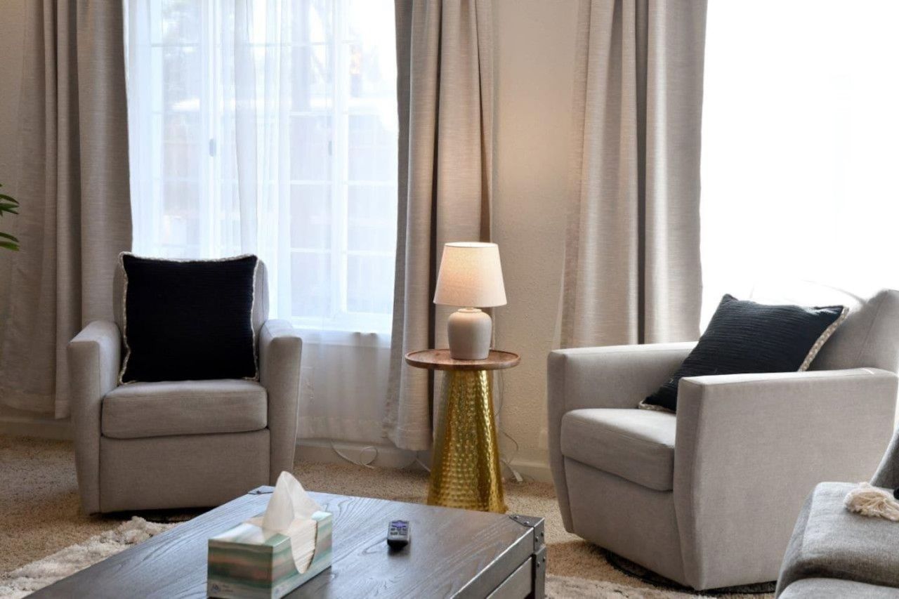 Una sala de estar con dos sillones, una mesa de centro, una lámpara y un ventanal con cortinas.