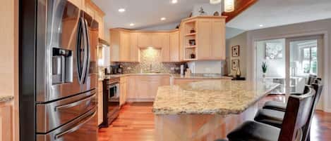 A modern kitchen with a stainless steel refrigerator, wooden cabinets, granite countertops, and a bar seating area.
