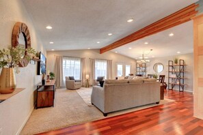 A spacious living room with a large sofa, a wooden ceiling beam, and a bookshelf.