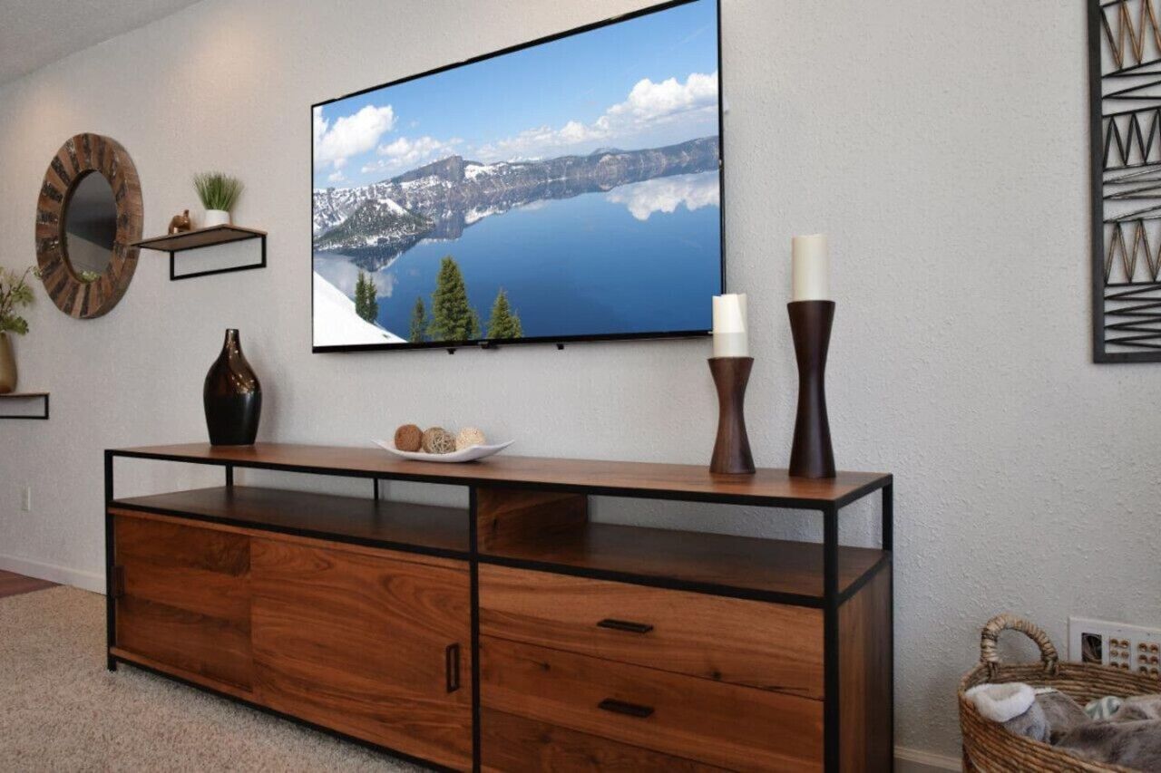 Un salon moderne avec un grand téléviseur à écran plat fixé au mur, une console en bois agrémentée d’objets décoratifs et un panier en osier posé sur le sol.