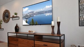 Ein modernes Wohnzimmer mit einem großen Flachbildfernseher an der Wand, einem Holzkonsolentisch mit Dekorationen und einem Korbwäschesammler auf dem Boden.