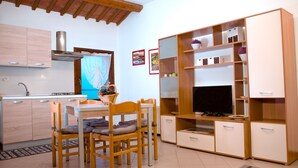 Cocina compacta con comedor, armarios de madera, televisión integrada y una ventana con cortinas translúcidas.