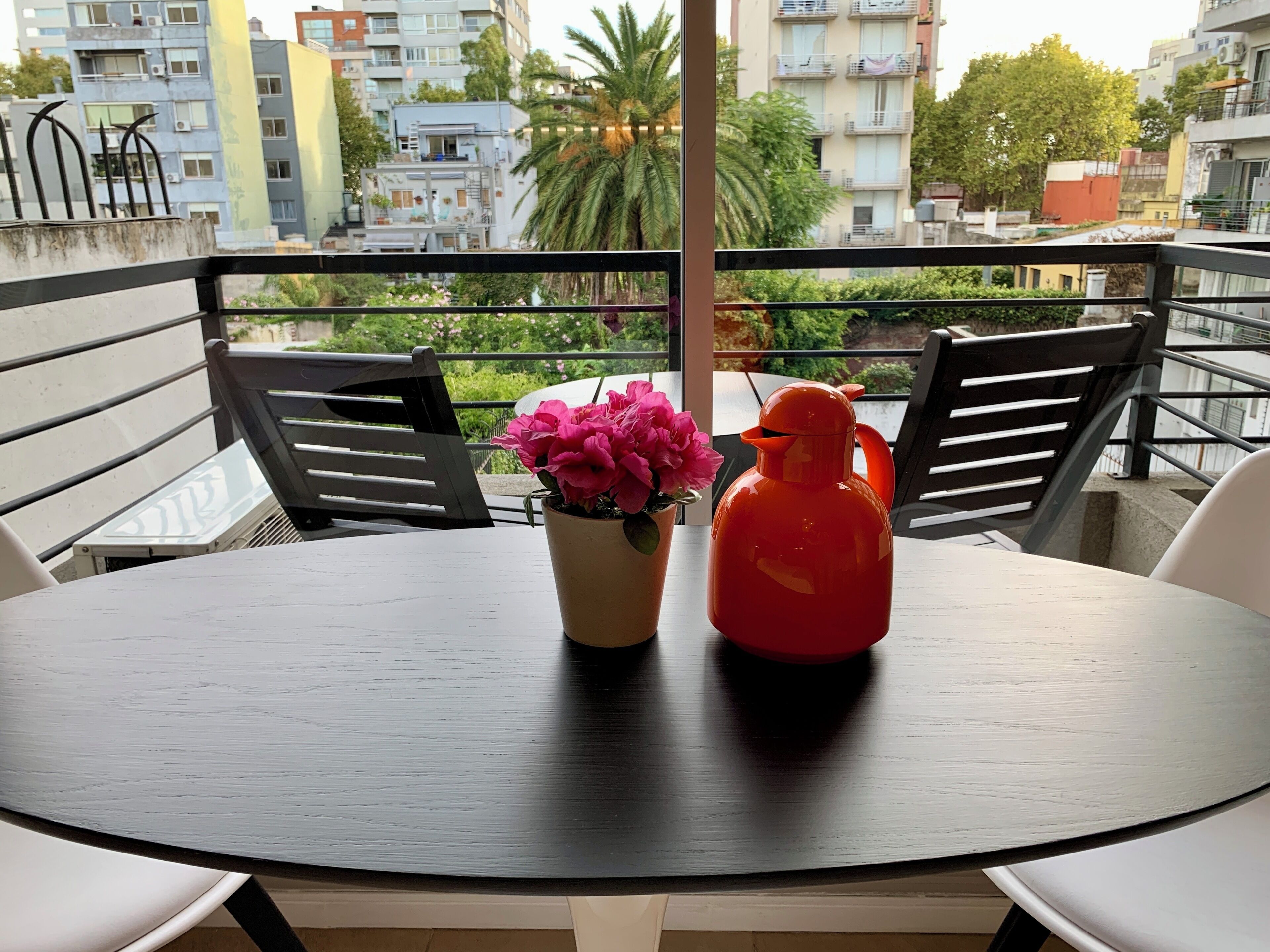Ein Balkon mit rundem Tisch, zwei Stühlen, einem Blumentopf mit rosa Blüten und einer roten Flasche.
