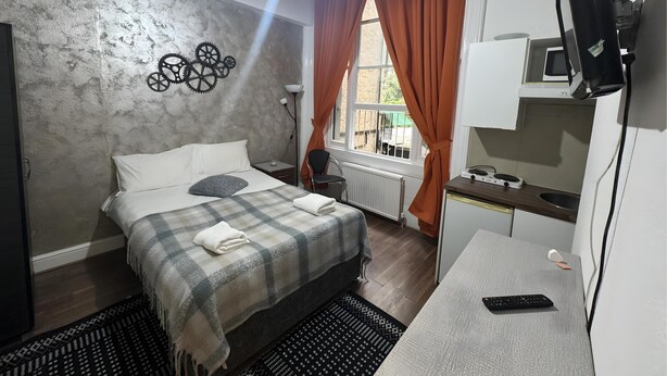A bedroom with a bed, a desk, a TV, and a window with curtains. - Studio Apartment Sleeps 2 with Ensuite (London)