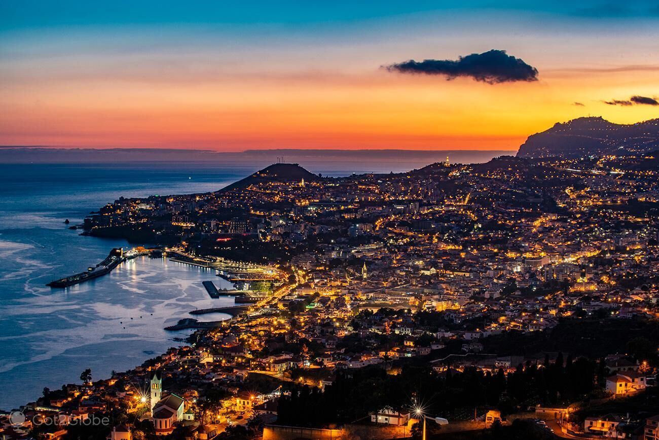 Une vue d’une ville côtière au crépuscule, avec une promenade en bord de mer bien visible et des bâtiments illuminés.