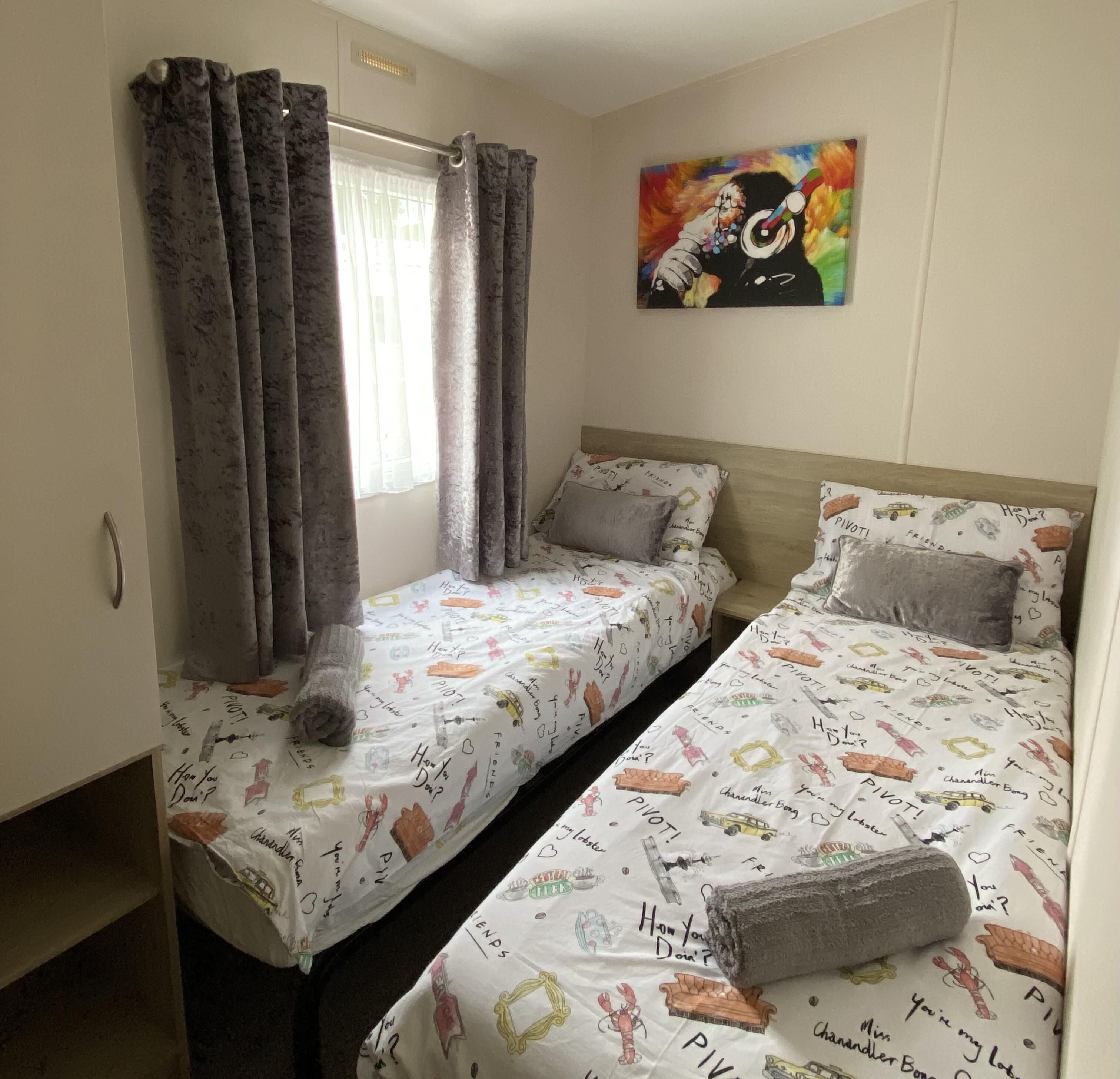 A room with two single beds, each with a patterned bedspread and pillows. There is a window with curtains, a painting on the wall, and a door on the left.