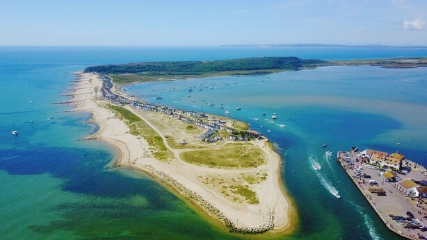 Vista aérea de uma área costeira com uma extensa praia de areia, uma pequena península com casas e uma marina com barcos.