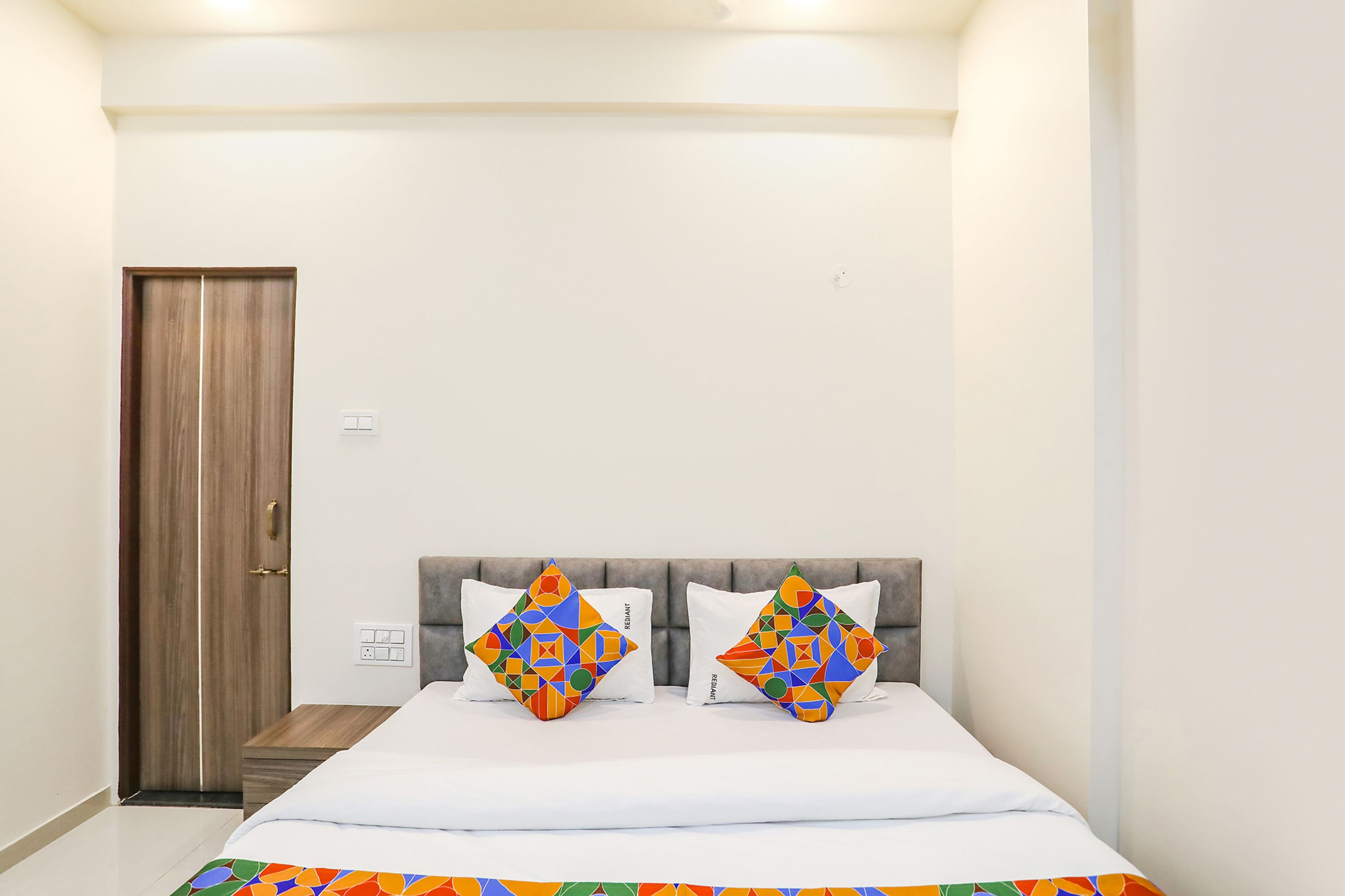 Une chambre à coucher avec un lit, deux coussins décoratifs, une porte en bois et un interrupteur sur le mur.