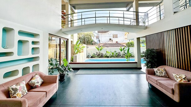 A spacious indoor area with a curved balcony, two sofas with patterned cushions, and a view of a swimming pool and greenery outside. - FabHotel Prime BT Wellness With Pool (Morjim)