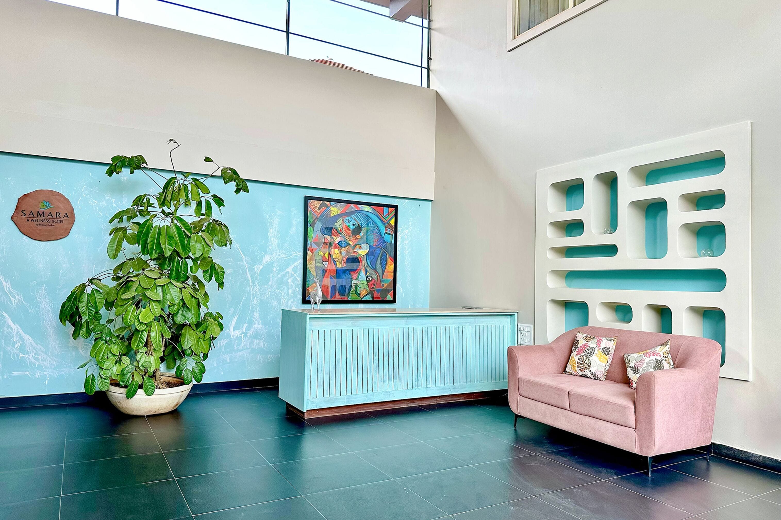 A reception area with a large potted plant, a blue reception desk, a pink sofa with patterned cushions, a colorful abstract painting, and a wall-mounted decorative panel.
