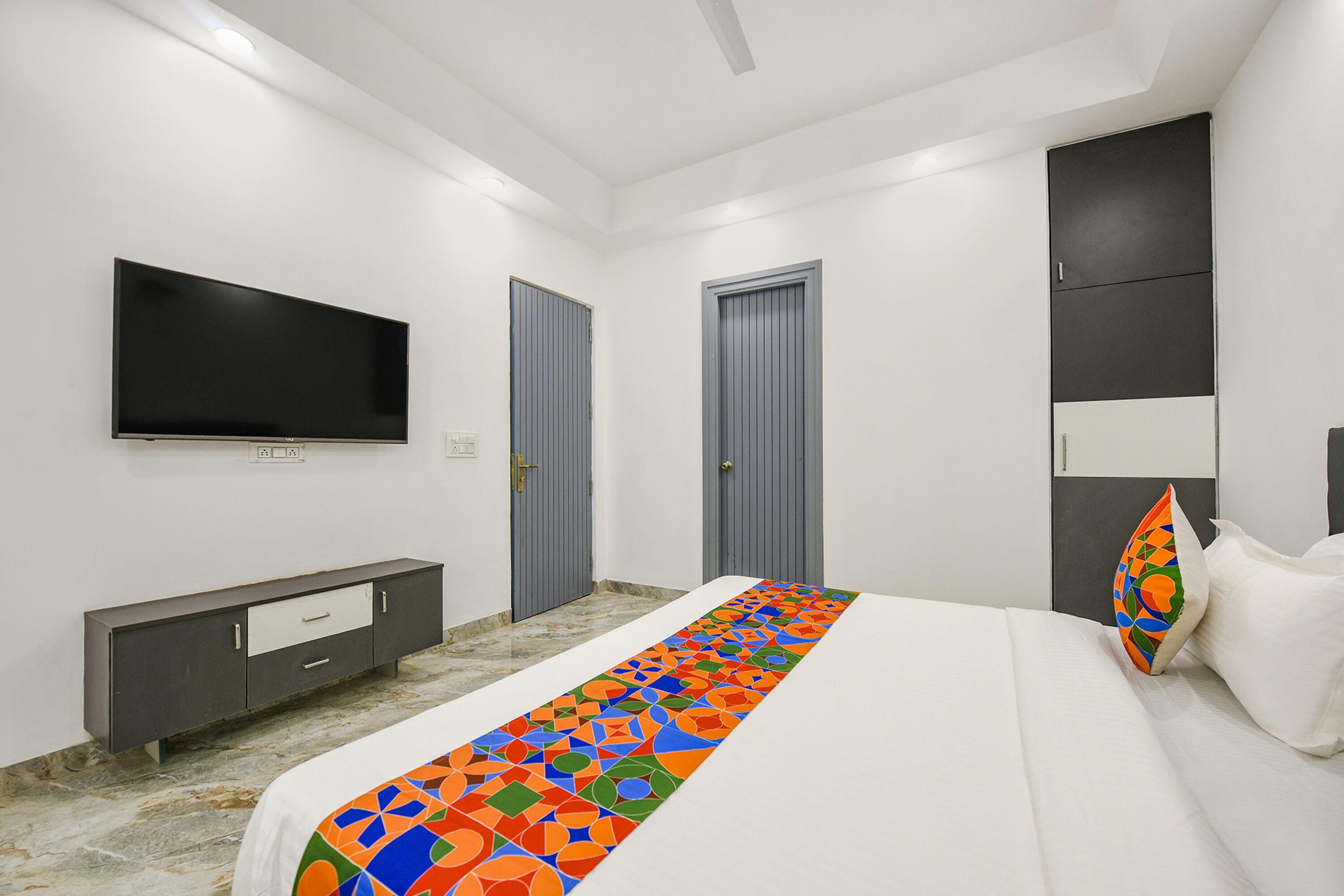 A modern bedroom with a large bed, a flat-screen TV mounted on the wall, a grey wardrobe, and a pair of doors.