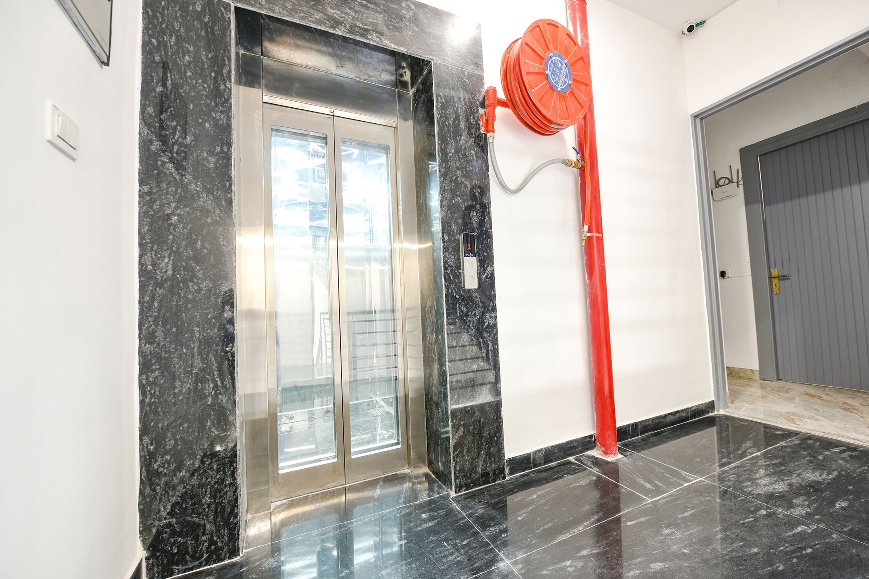 An elevator with a glass door, a red fire hose, and a grey door to the right.