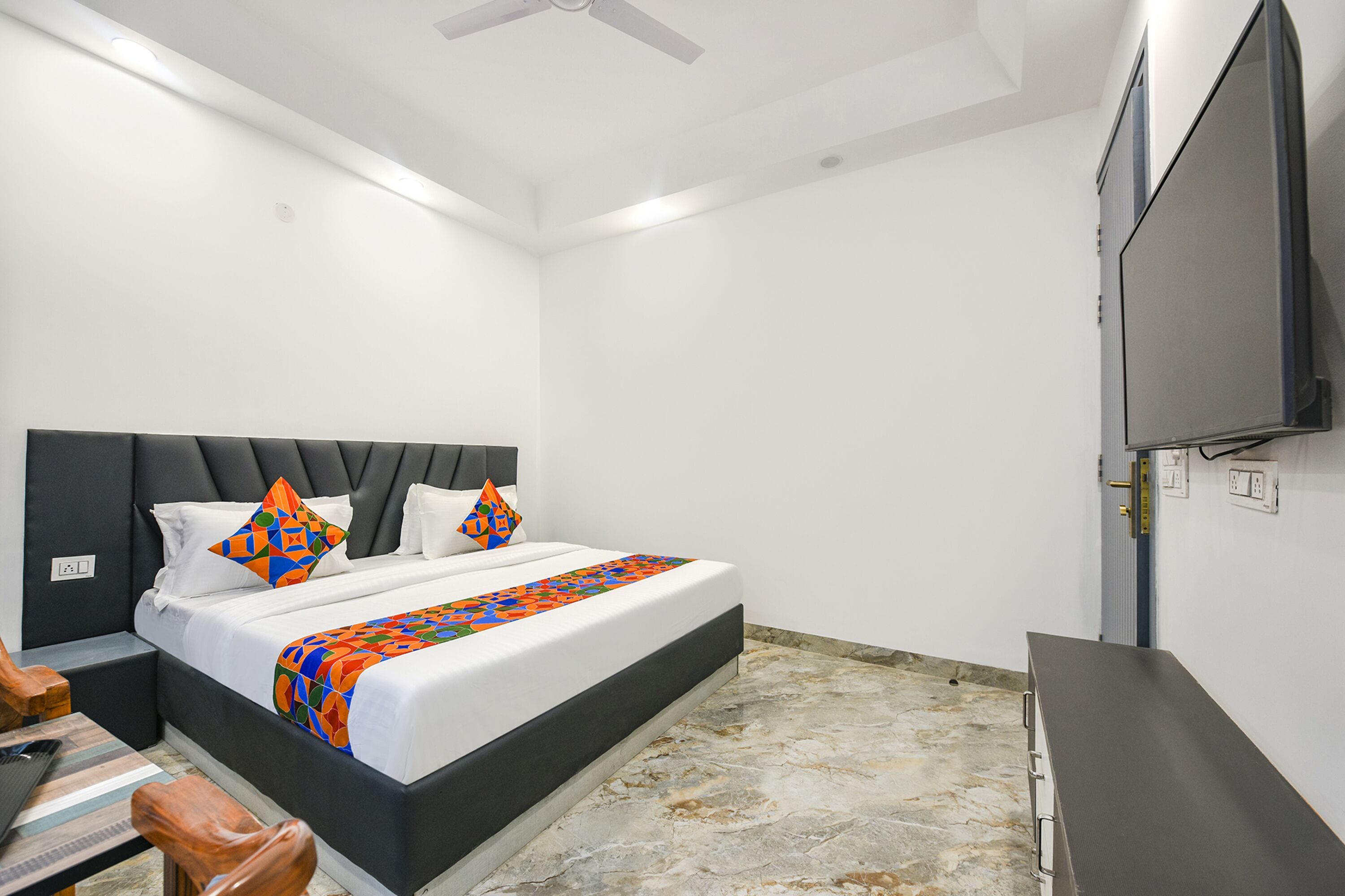 A bedroom with a bed, a flat-screen TV, a ceiling fan, and a stone floor.
