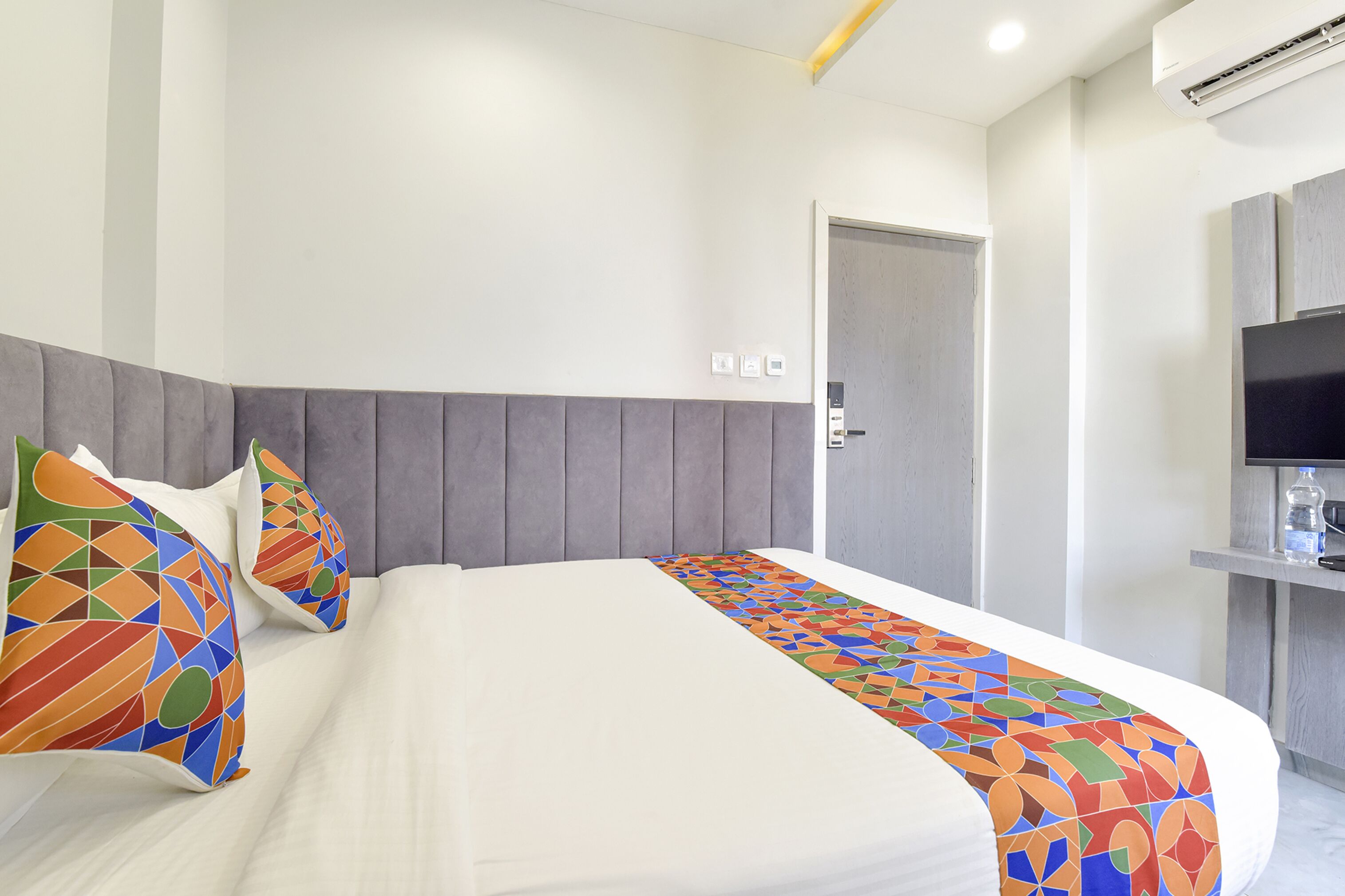 A hotel room with a bed, a gray headboard, a TV, and a door.