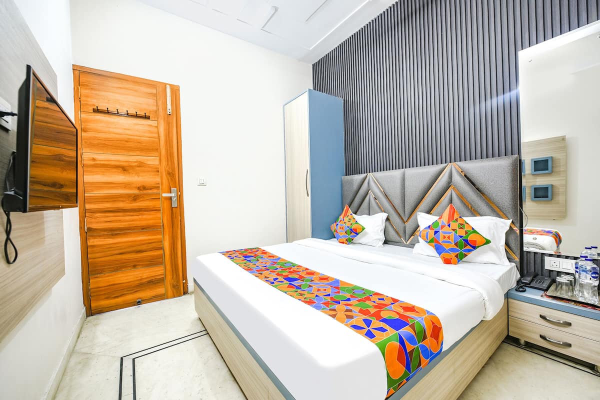 A modern hotel room with a wooden door, a bed with a colorful geometric bedspread, a headboard with a geometric pattern, a bedside table with a phone and a water bottle, and a mirror on the wall.