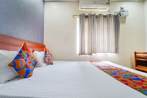 Une chambre d’hôtel avec un grand lit, des oreillers colorés, une longue table, une chaise en bois, un climatiseur et une fenêtre avec des rideaux.