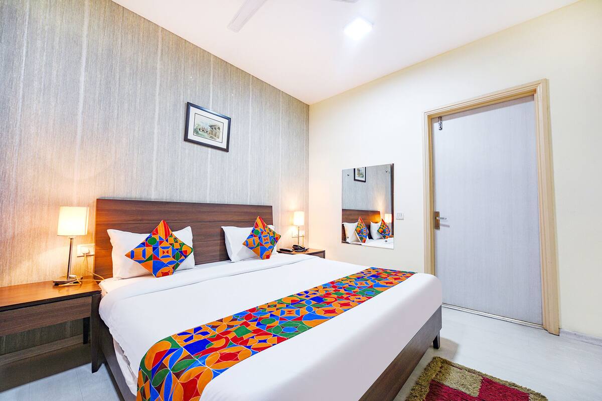 A hotel room with a large bed, two bedside tables with lamps, a wooden headboard, a colorful bedspread, a framed picture on the wall, a mirror, and a door.