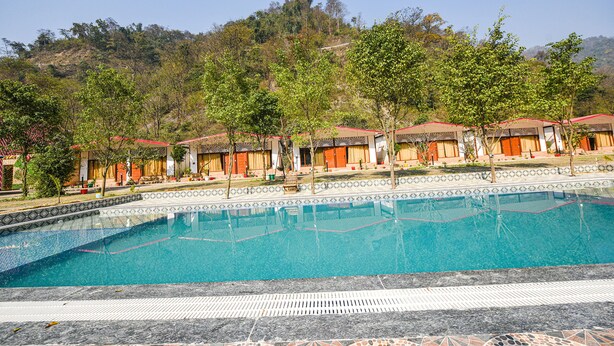 A swimming pool with tiled edges, surrounded by a row of buildings with red roofs and beige walls, set against a backdrop of a hilly landscape.