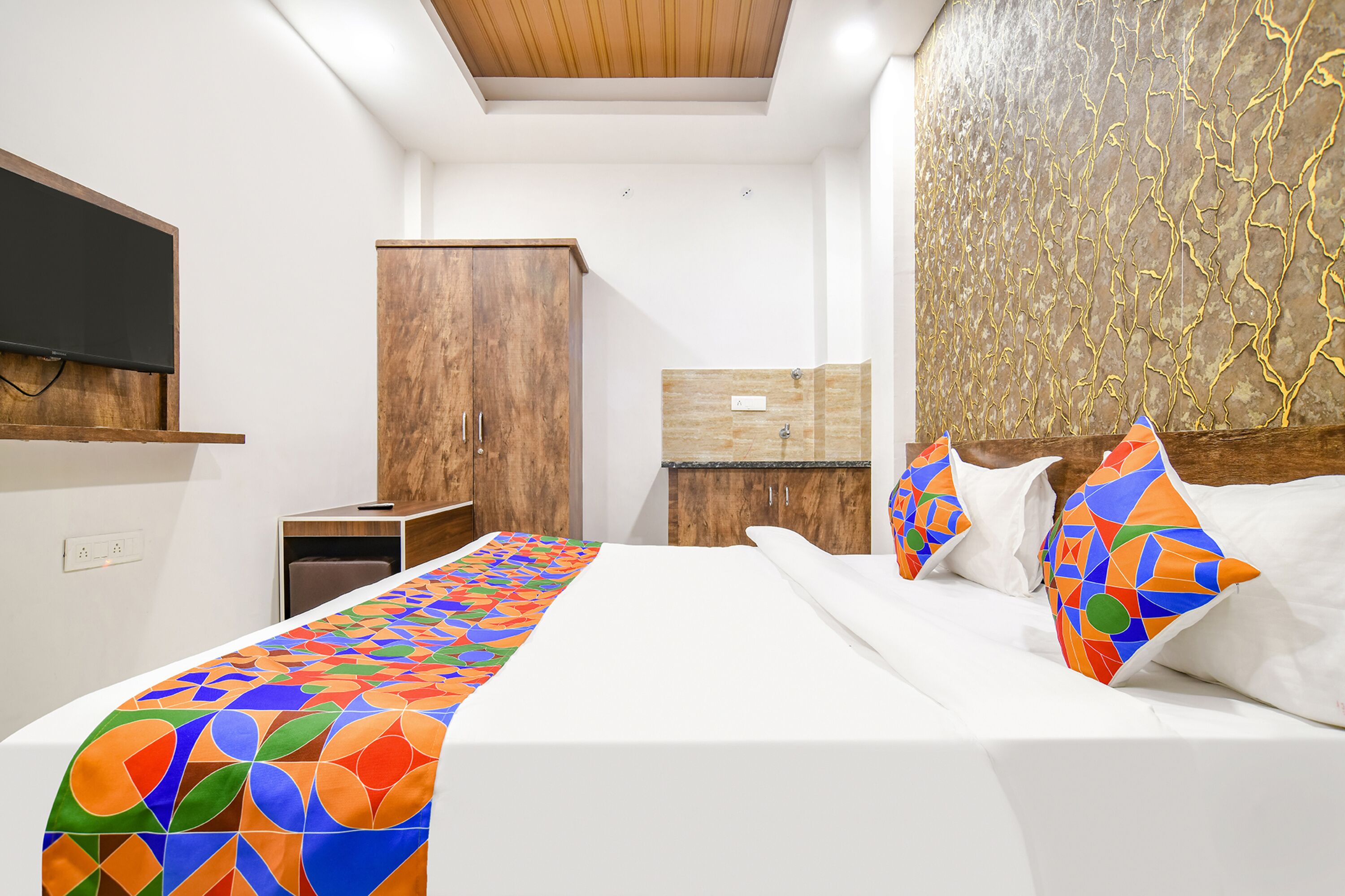 A modern hotel room with a large bed, a wooden wardrobe, a TV mounted on the wall, and a patterned headboard.
