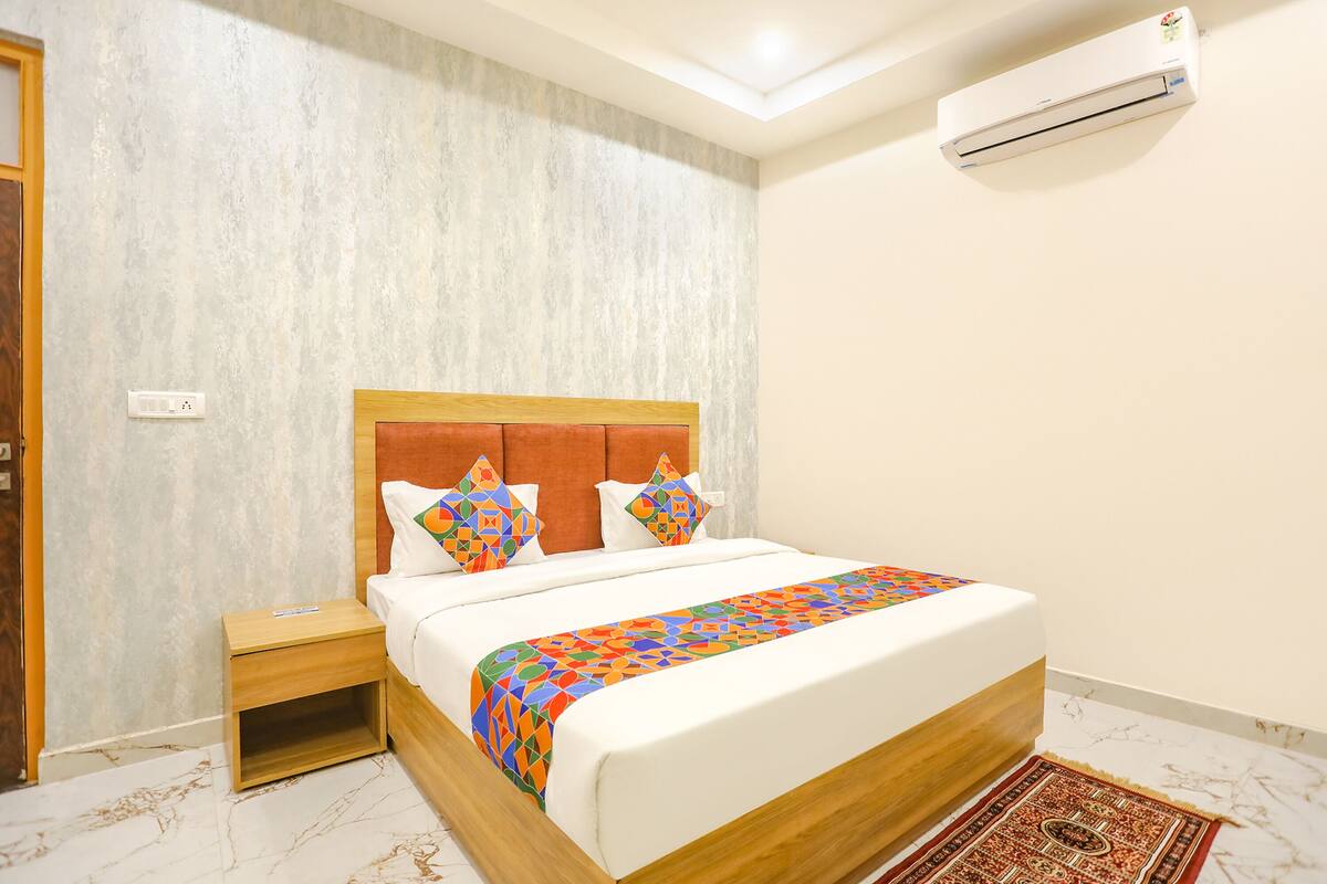 Uma cama bem arrumada com colcha estampada, uma mesinha de cabeceira de madeira, um chão com padrão de mármore e um ar condicionado fixado na parede.