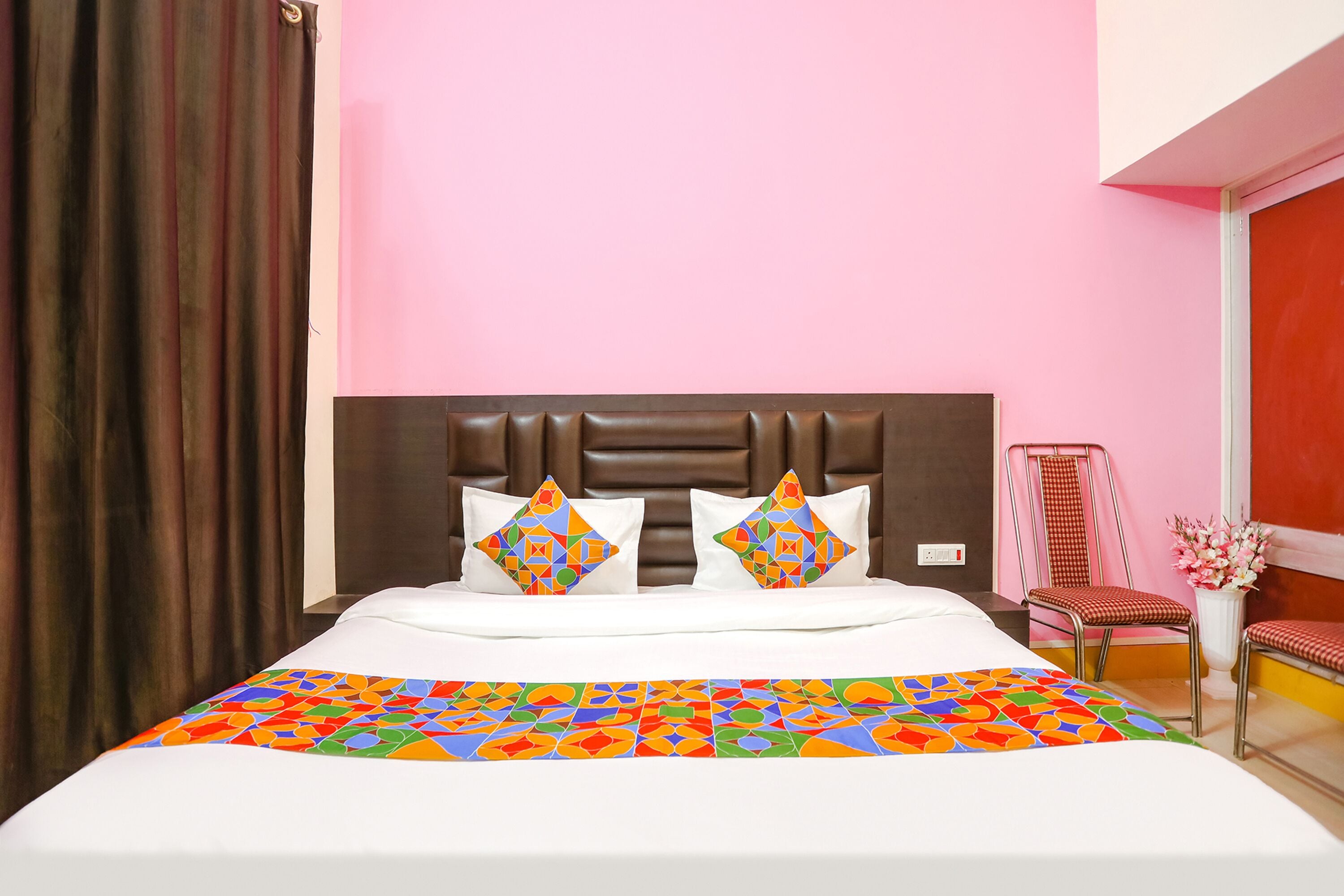A hotel room with a bed, two decorative pillows, a colorful bedspread, a headboard, a chair, and a vase with flowers.