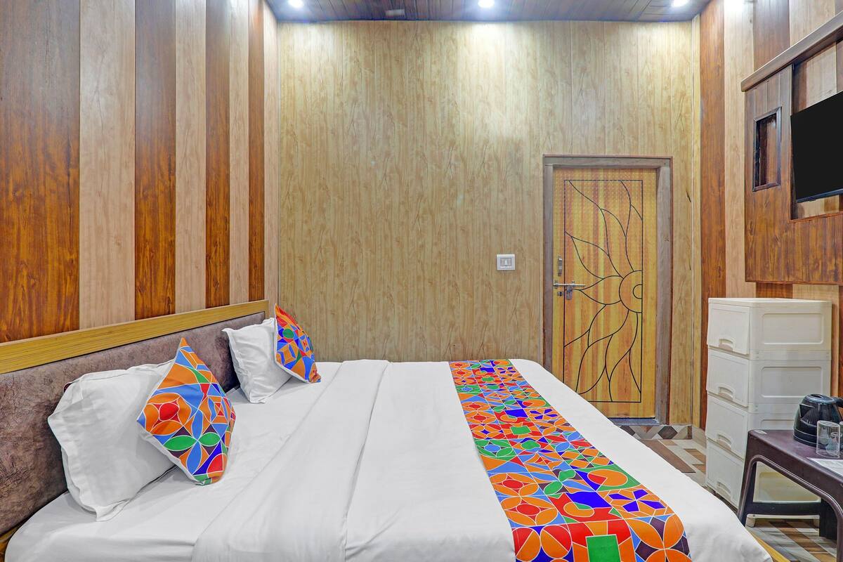 A hotel room with a wooden paneled wall, a bed with white bedding and colorful pillows, a door with a geometric design, and a small white cabinet.