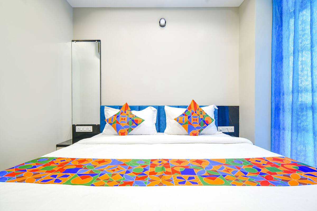 A hotel room with a bed, two pillows, a nightstand, a lamp, and a window with blue curtains.