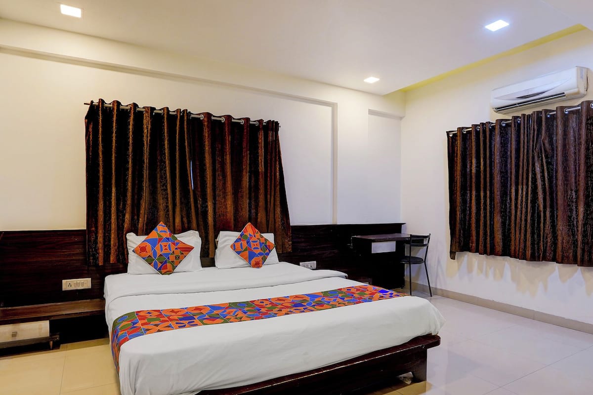 A hotel room with a large bed, two pillows with colorful geometric patterns, dark curtains, a bedside table, a desk, and an air conditioning unit.