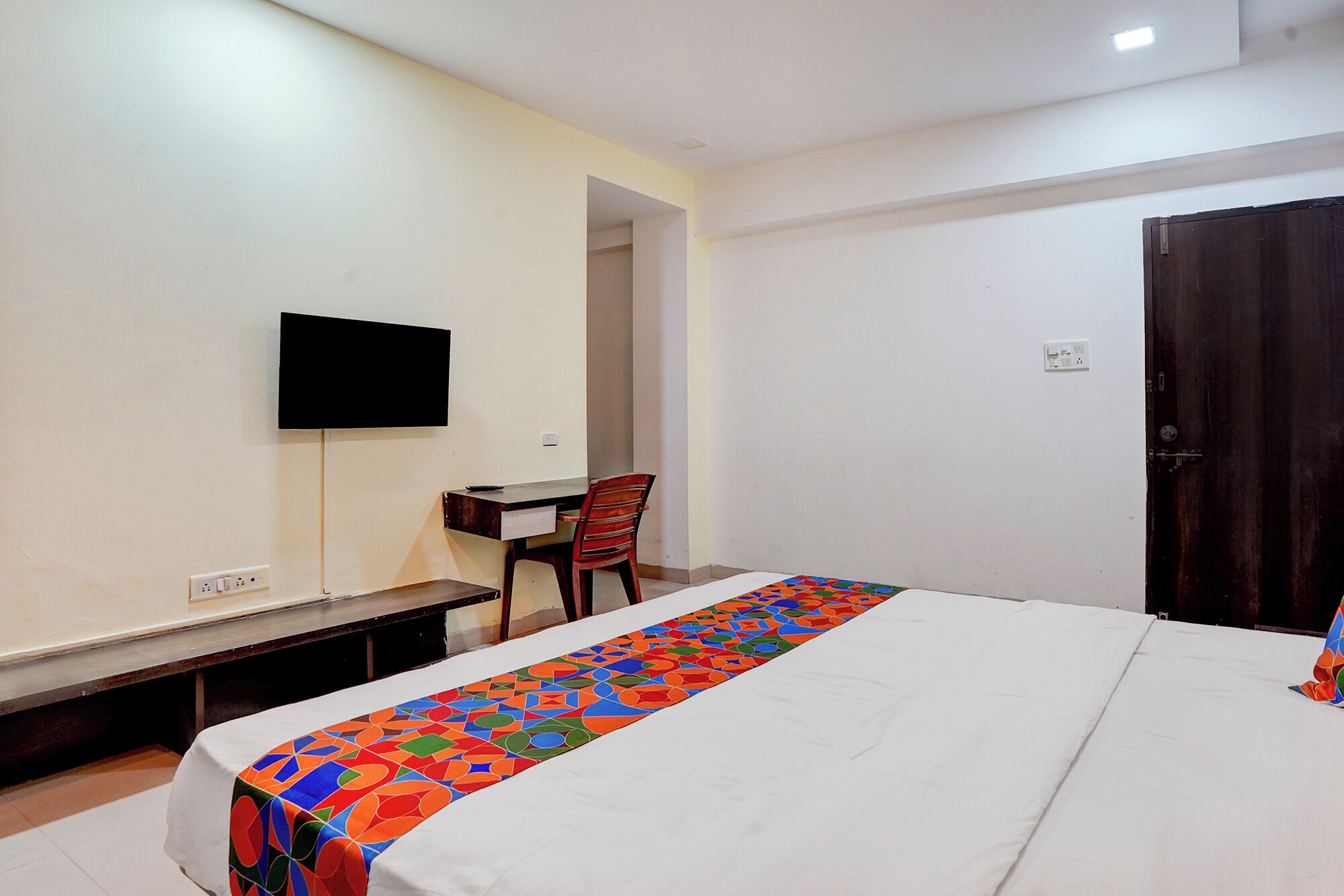 A hotel room with a bed, a desk, a chair, a television mounted on the wall, and a door.