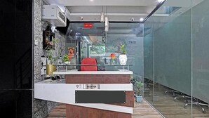 A reception desk with a glass partition, a red chair, and potted plants. - Fabhotel Bellmore Stay & Homes (Gurugram)