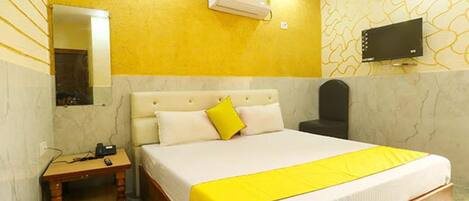 A hotel room with a bed, a bedside table, a chair, a TV, and an air conditioning unit.