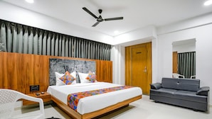 A modern hotel room with a wooden headboard, a bed with colorful bedding, a grey sofa, a white chair, and a ceiling fan.