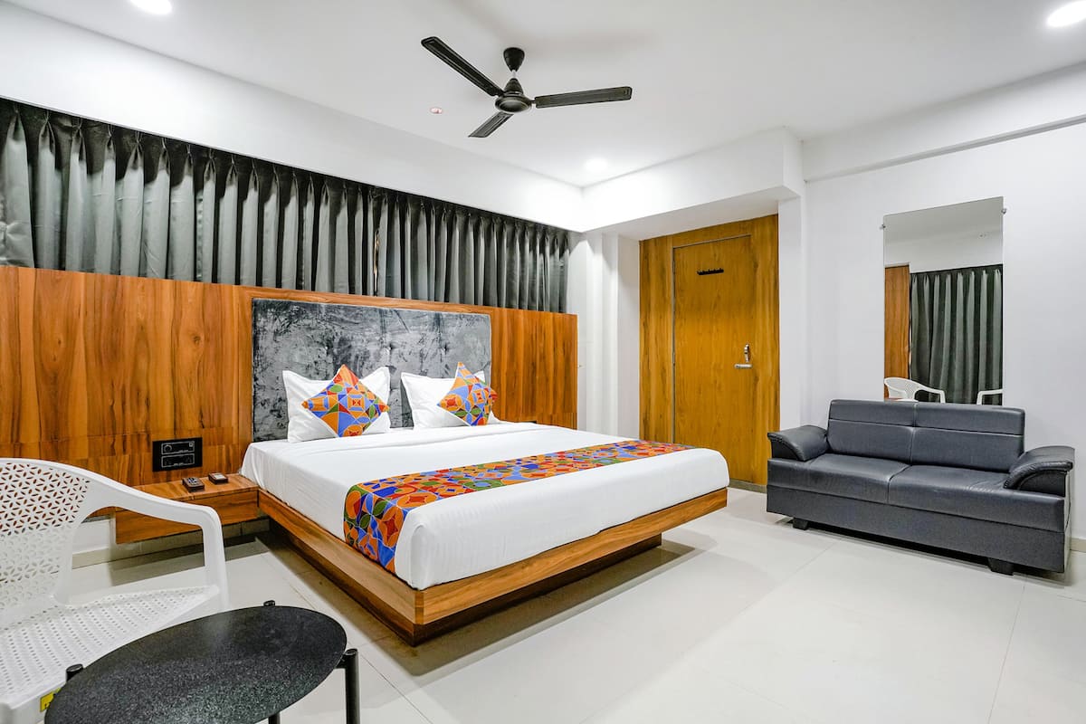Una habitación de hotel moderna con cabecera de madera, cama con ropa de cama colorida, sofá gris, silla blanca y ventilador de techo.