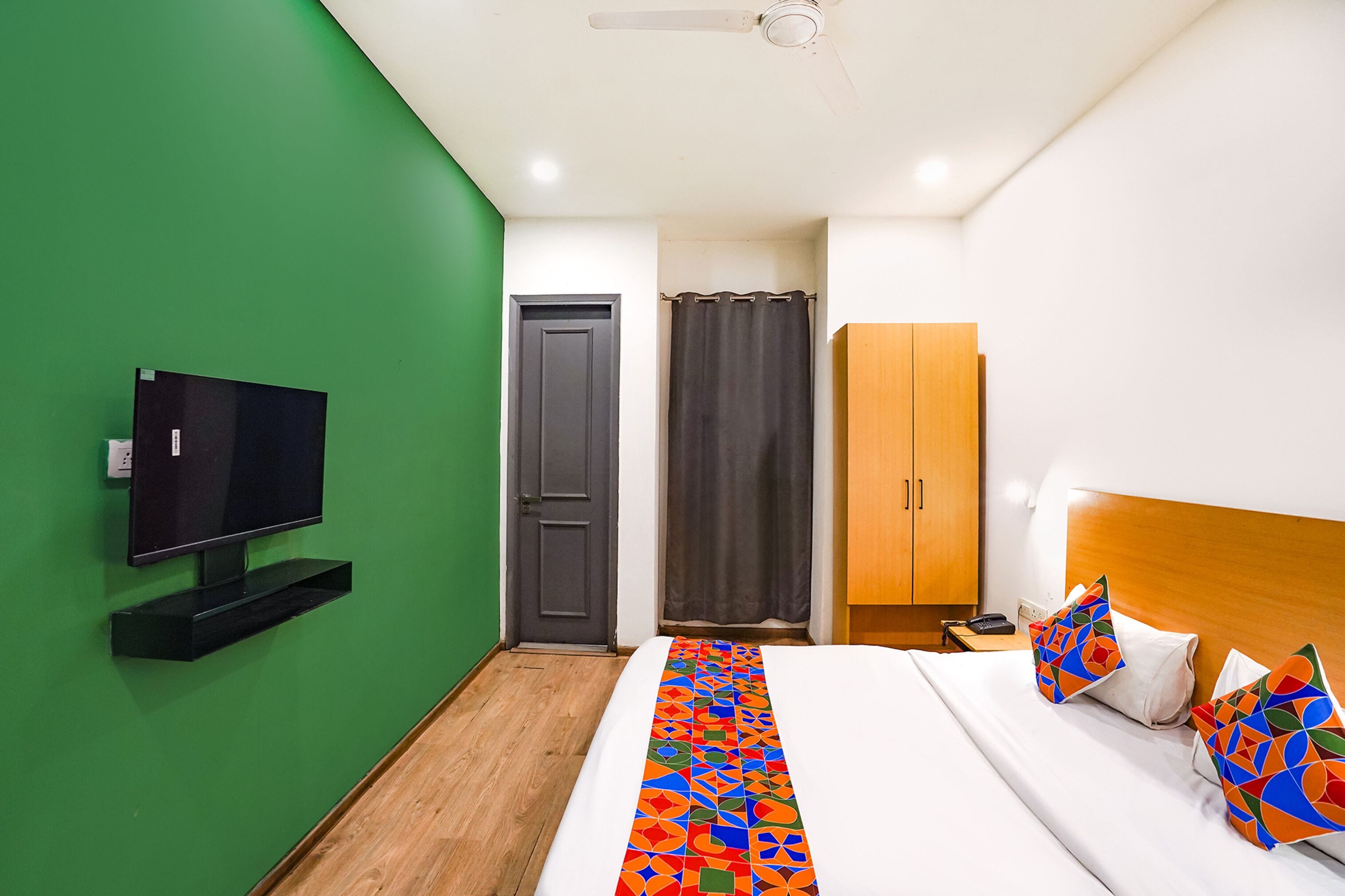 A hotel room with a green wall, a flat-screen TV mounted on the wall, a bed with colorful pillows, a wooden wardrobe, and a ceiling fan.