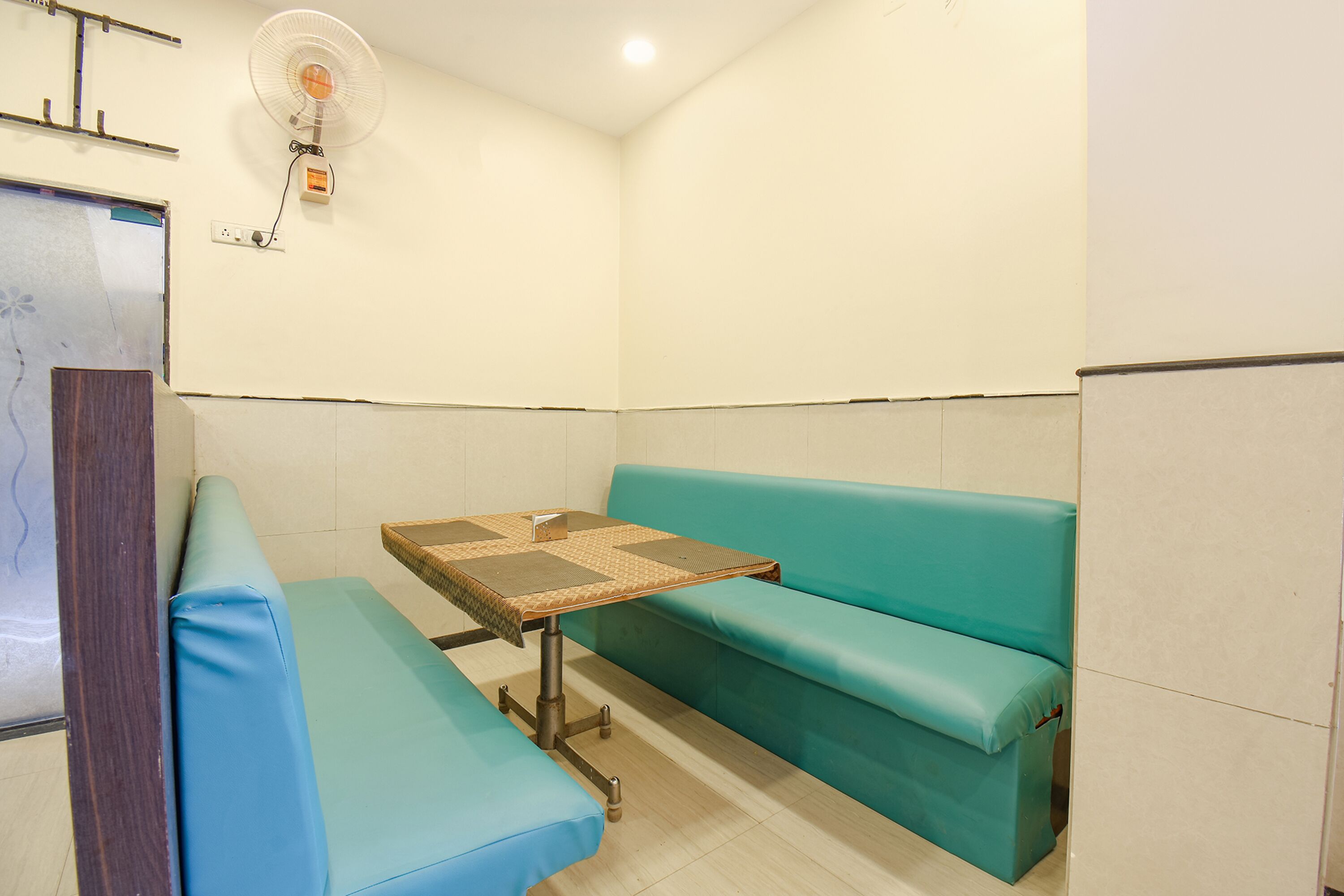 A small room with a teal-colored bench, a small table with a woven top, and a wall-mounted fan.