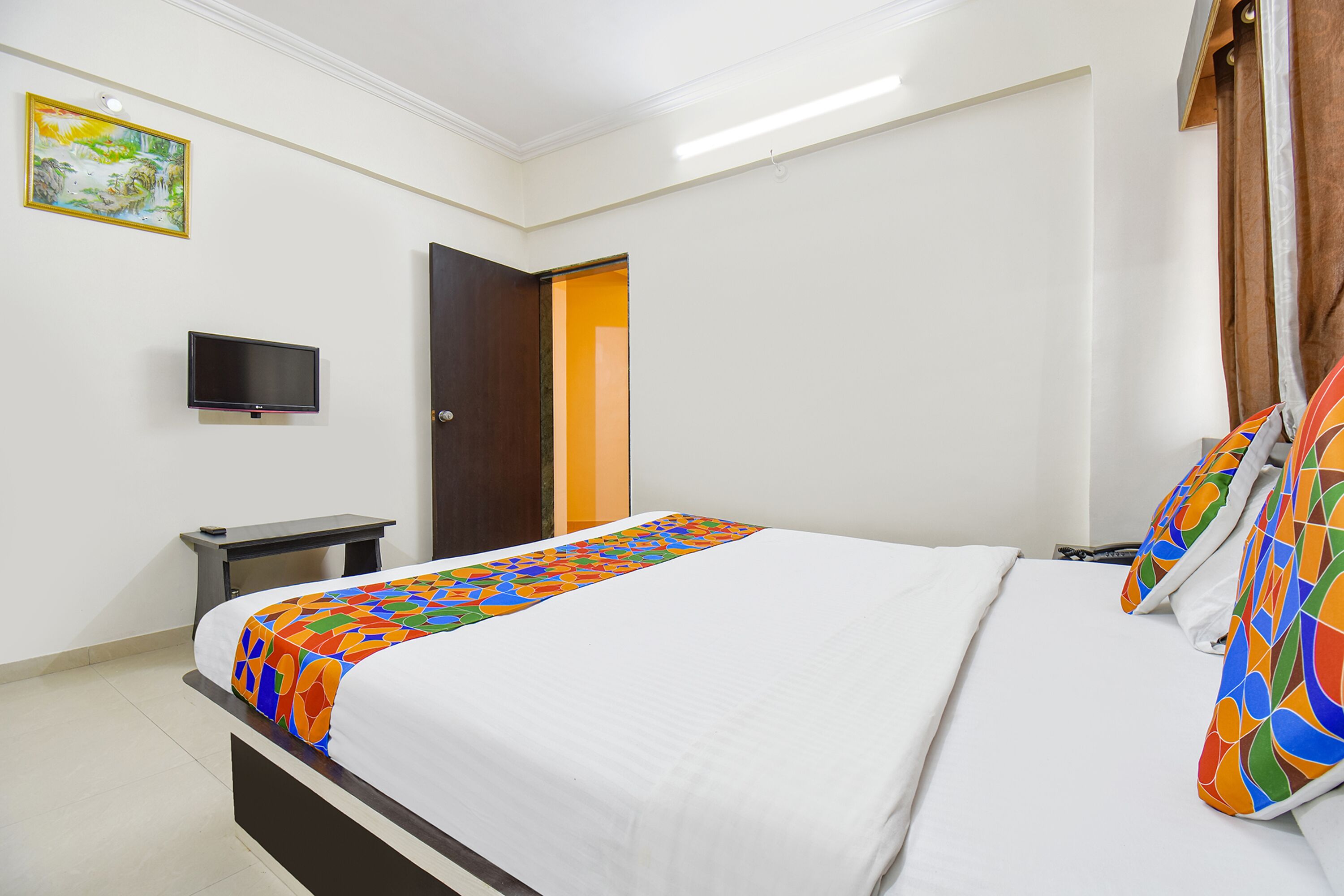 Una cama bien hecha con un edredón colorido, un televisor de pantalla plana sobre un mueble, y una puerta que da a otra habitación.