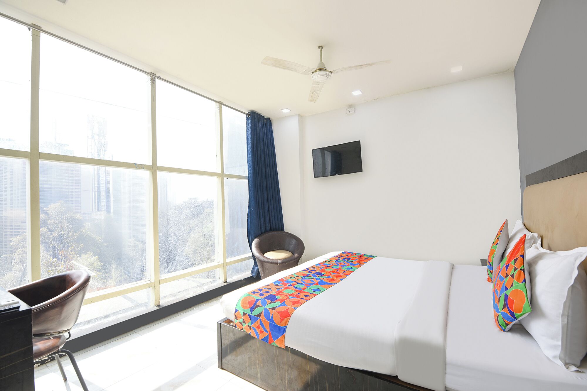 A hotel room with a large window, a bed with colorful bedding, a chair, and a ceiling fan.