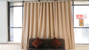Ett rum med ett stort fönster, beige gardiner, en mörk soffa med två bruna kuddar och en skylt med texten ”Rökning förbjuden” på fönstret.