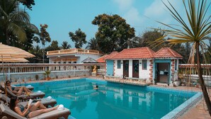 A swimming pool with lounge chairs and umbrellas, surrounded by buildings and trees. - FabHotel Prime Indeedcare (Kakdwip)
